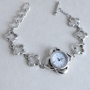 May include: Silver-toned bracelet watch with a white watch face. The bracelet features a clover-shaped design with small, clear crystals. The watch face is round with black hands and hour markers.