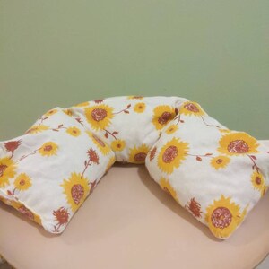 May include: Two white cushions with a sunflower pattern on a pink table. The cushions are shaped like a crescent moon and are made of a soft, comfortable fabric.