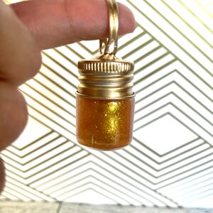 Op de afbeelding: Een klein gouden flesje met een gouden dop en een gouden ketting die aan de dop is bevestigd. Het flesje is gevuld met gouden glitter.
