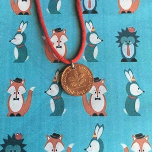 May include: A red cord necklace with a gold-colored coin pendant. The coin features a leaf design and the text "BUNDESREPUBLIK DEUTSCHLAND 1986". The background is a light blue fabric with repeating cartoon fox, rabbit, and hedgehog illustrations.
