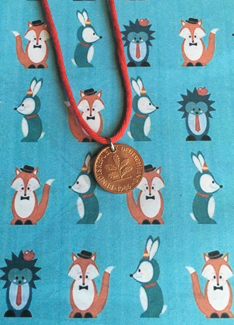 May include: A red cord necklace with a gold-colored coin pendant. The coin features a leaf design and the text "BUNDESREPUBLIK DEUTSCHLAND 1986". The background is a light blue fabric with repeating cartoon fox, rabbit, and hedgehog illustrations.