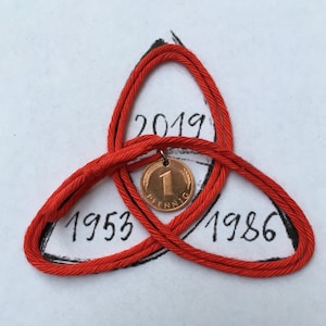May include: A red cord knot with a copper coin pendant. The coin has the number 1 and the word "Pfennig" on it. The knot is drawn over the numbers 1953, 1986, and 2019.