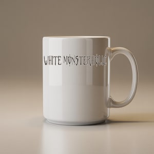 May include: A white ceramic mug with a clear handle. The mug has the text "WHITE MONSTERHOLIC" in a black, stylized font. The cylindrical mug is on a light-coloured surface.