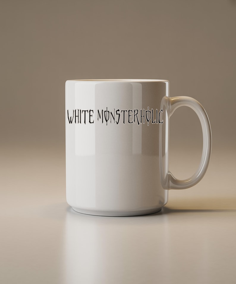 May include: A white ceramic mug with a clear handle. The mug has the text "WHITE MONSTERHOLIC" in a black, stylized font. The cylindrical mug is on a light-coloured surface.