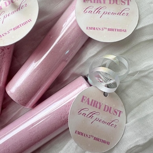 May include: Clear plastic tubes filled with pink bath powder, each topped with a white ribbon and a round tag printed with the text FAIRY DUST bath powder and EMMA'S 5TH BIRTHDAY in pink lettering, creating a set of party favors.