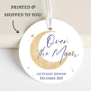 May include: A round white tag with a gold glitter crescent moon and navy blue script that reads "Over the Moon." The tag includes the text "Liza's Baby Shower December 2025" and is strung with white cord.