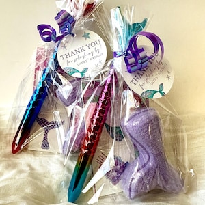 May include: Two cellophane gift bags tied with purple ribbon, each containing a mermaid-themed pen, a mermaid tail-shaped item, and a tag that reads "THANK YOU for splashing by!" for Lily's 5th Birthday.