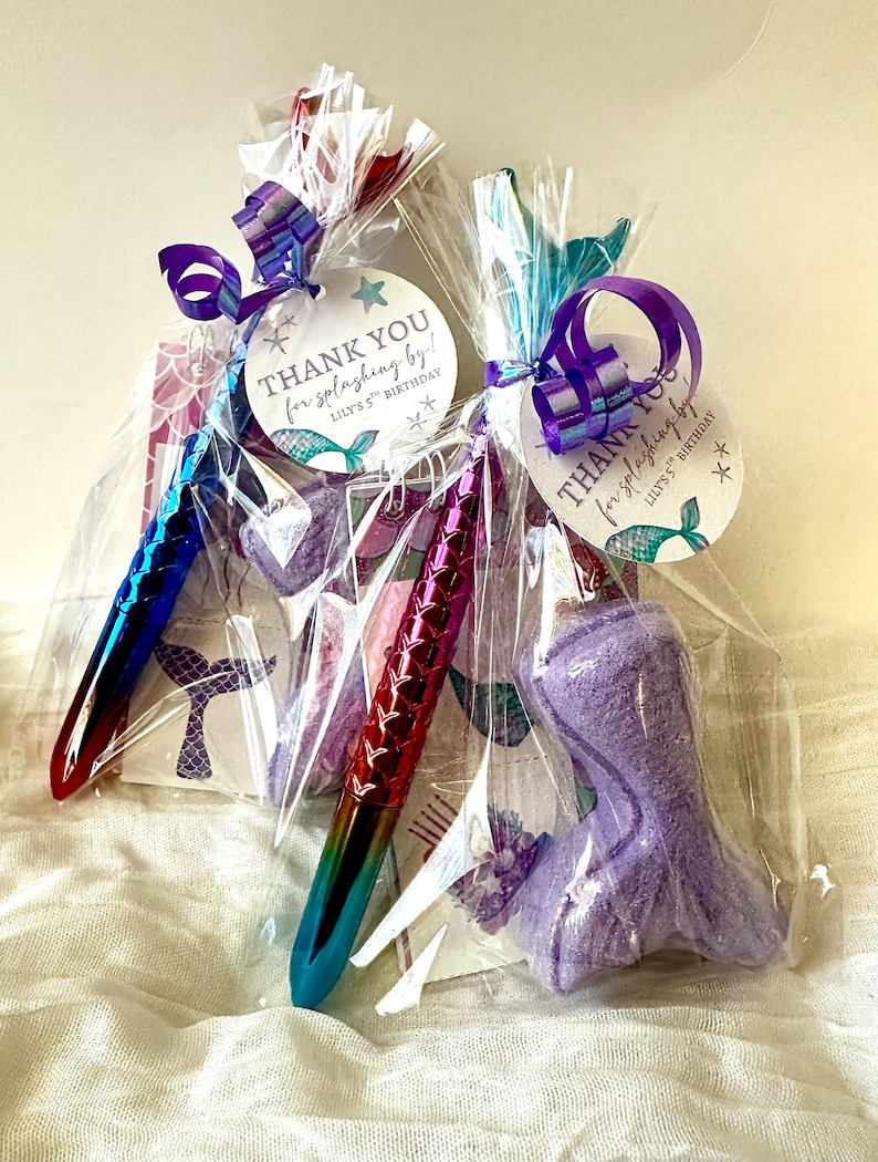 May include: Two cellophane gift bags tied with purple ribbon, each containing a mermaid-themed pen, a mermaid tail-shaped item, and a tag that reads "THANK YOU for splashing by!" for Lily's 5th Birthday.