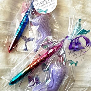 May include: Two party favors wrapped in clear cellophane. Each includes a mermaid-themed pen with a color-shifting finish and a purple mermaid tail-shaped bath bomb. The tags read "THANK YOU for splashing by!" for a 5th birthday.