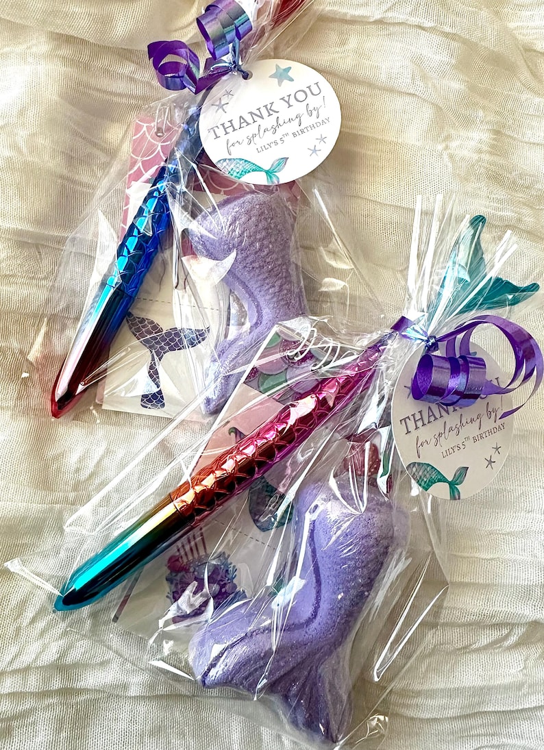 May include: Two party favors wrapped in clear cellophane. Each includes a mermaid-themed pen with a color-shifting finish and a purple mermaid tail-shaped bath bomb. The tags read "THANK YOU for splashing by!" for a 5th birthday.