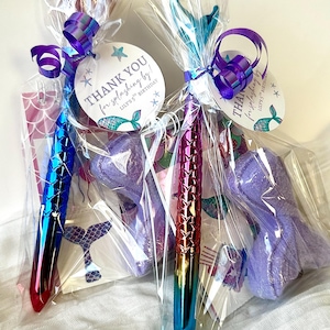 May include: Two mermaid-themed party favors in clear cellophane bags. Each bag contains a pen with a scale design, a purple bath bomb, and a tag that reads "THANK YOU for splashing by!" with a mermaid tail illustration. The pens have a gradient color scheme.