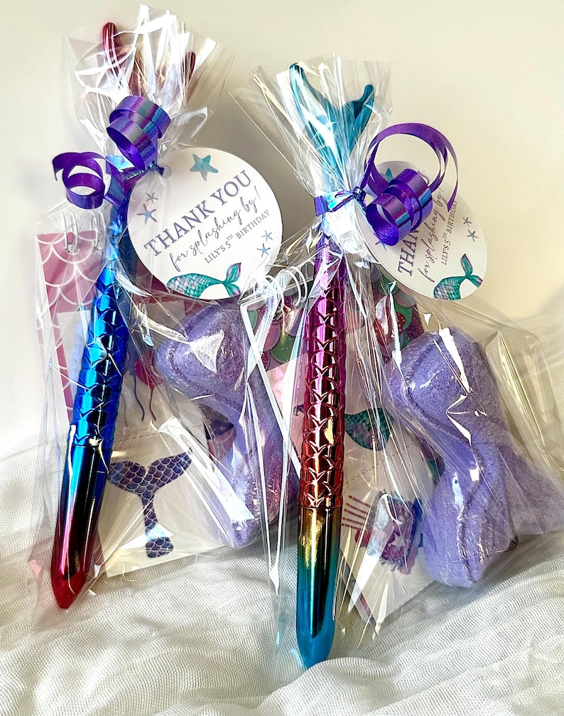 May include: Two mermaid-themed party favors in clear cellophane bags. Each bag contains a pen with a scale design, a purple bath bomb, and a tag that reads "THANK YOU for splashing by!" with a mermaid tail illustration. The pens have a gradient color scheme.