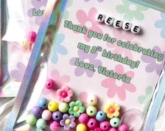 Puede incluir: Una bolsa holográfica resellable que contiene cuentas de plástico de colores y una tarjeta con estampado floral. La tarjeta muestra el nombre REESE en cuentas de letras blancas y el texto: Thank you for celebrating my 8th birthday! Love, Victoria.
