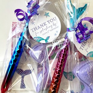 May include: Two mermaid-themed party favors in clear cellophane bags, each tied with a purple ribbon. One pen is blue and red, the other is red and gold. Each favor has a tag that says "THANK YOU for splashing by!" and "Lily's 5th Birthday".