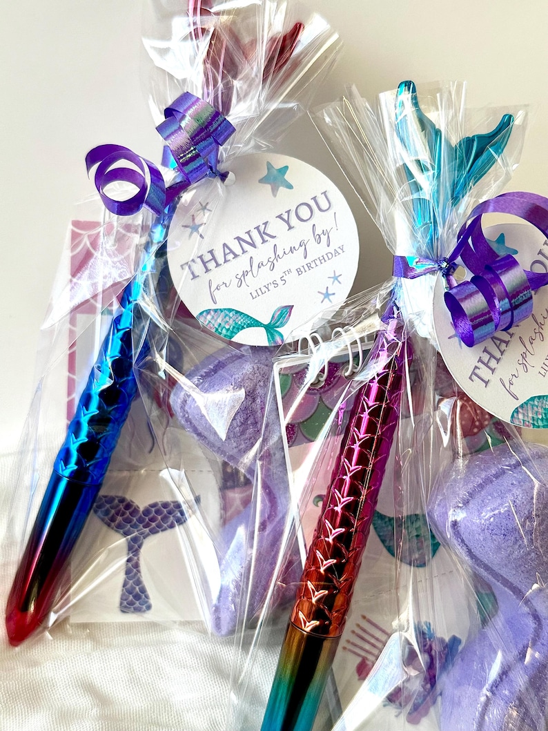 May include: Two mermaid-themed party favors in clear cellophane bags, each tied with a purple ribbon. One pen is blue and red, the other is red and gold. Each favor has a tag that says "THANK YOU for splashing by!" and "Lily's 5th Birthday".