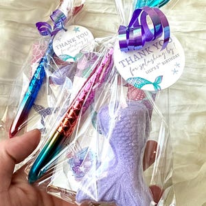 May include: Two clear cellophane bags containing mermaid-themed party favors. One bag has a purple mermaid tail-shaped item and a pen with a blue to green gradient. The other bag has a pen with a red to pink gradient. Each bag has a tag that says "THANK YOU for splashing by!"