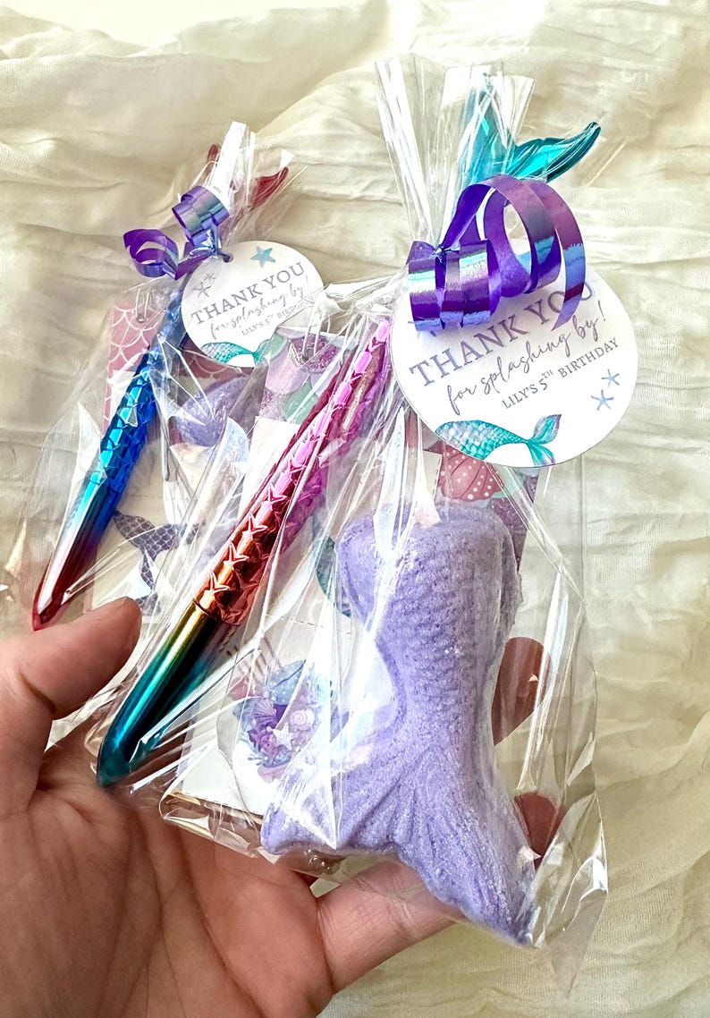 May include: Two clear cellophane bags containing mermaid-themed party favors. One bag has a purple mermaid tail-shaped item and a pen with a blue to green gradient. The other bag has a pen with a red to pink gradient. Each bag has a tag that says "THANK YOU for splashing by!"