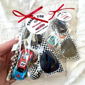 May include: Two clear plastic favor bags containing checkered sunglasses, a blue and red toy race car with the number 32, a checkered pencil, and a checkered ball. Each bag has a red ribbon and a tag reading THANK YOU FOR RACING BY ETHAN'S 8TH BIRTHDAY.