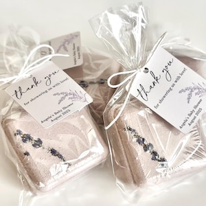 May include: Two square, light pink bath bombs, decorated with lavender sprigs, are packaged in clear cellophane. Each is tied with a white ribbon and a tag that reads "thank you for showering us with love!" and "Angela's Baby Shower August 2025."
