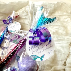 May include: Gift bags with mermaid-themed party favors. Each bag contains a purple bath bomb, a pen, and a tag that reads "Thank you for splashing by!" with a mermaid tail illustration. The bags are tied with purple and teal ribbons.