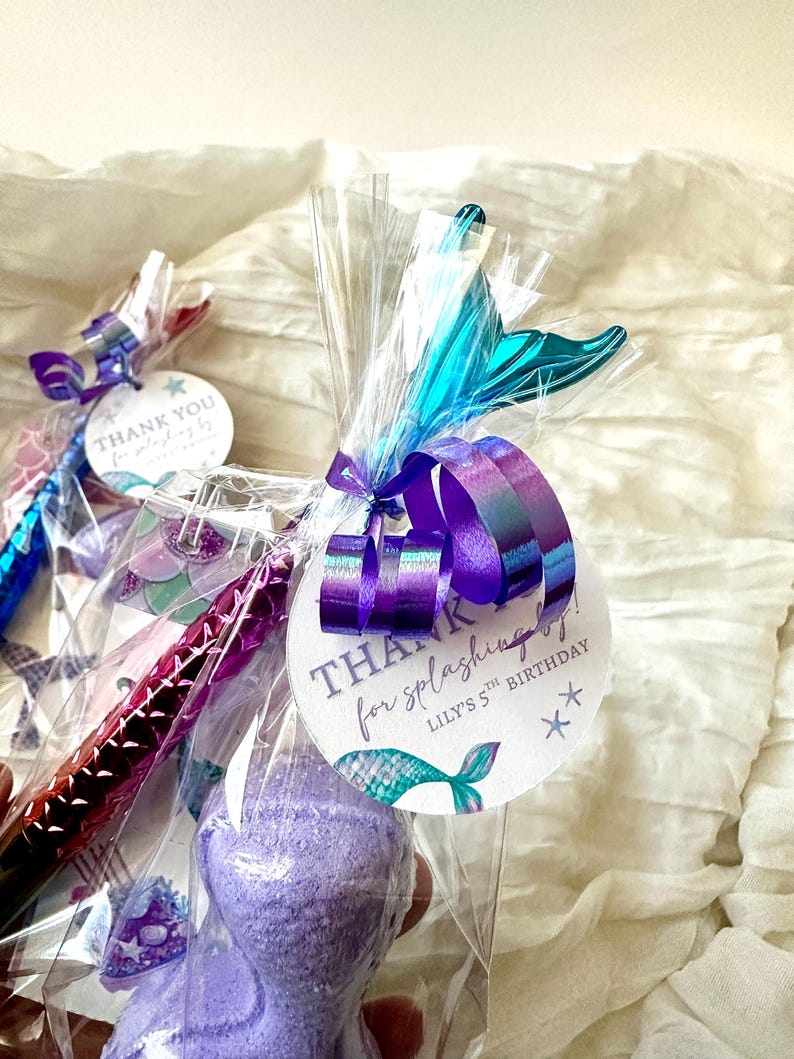 May include: Gift bags with mermaid-themed party favors. Each bag contains a purple bath bomb, a pen, and a tag that reads "Thank you for splashing by!" with a mermaid tail illustration. The bags are tied with purple and teal ribbons.