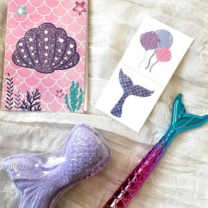 May include: A collection of mermaid-themed items, including a pink spiral notebook with a seashell design, temporary tattoos with balloons and a mermaid tail, a purple bath bomb, and a pen with a mermaid tail design.