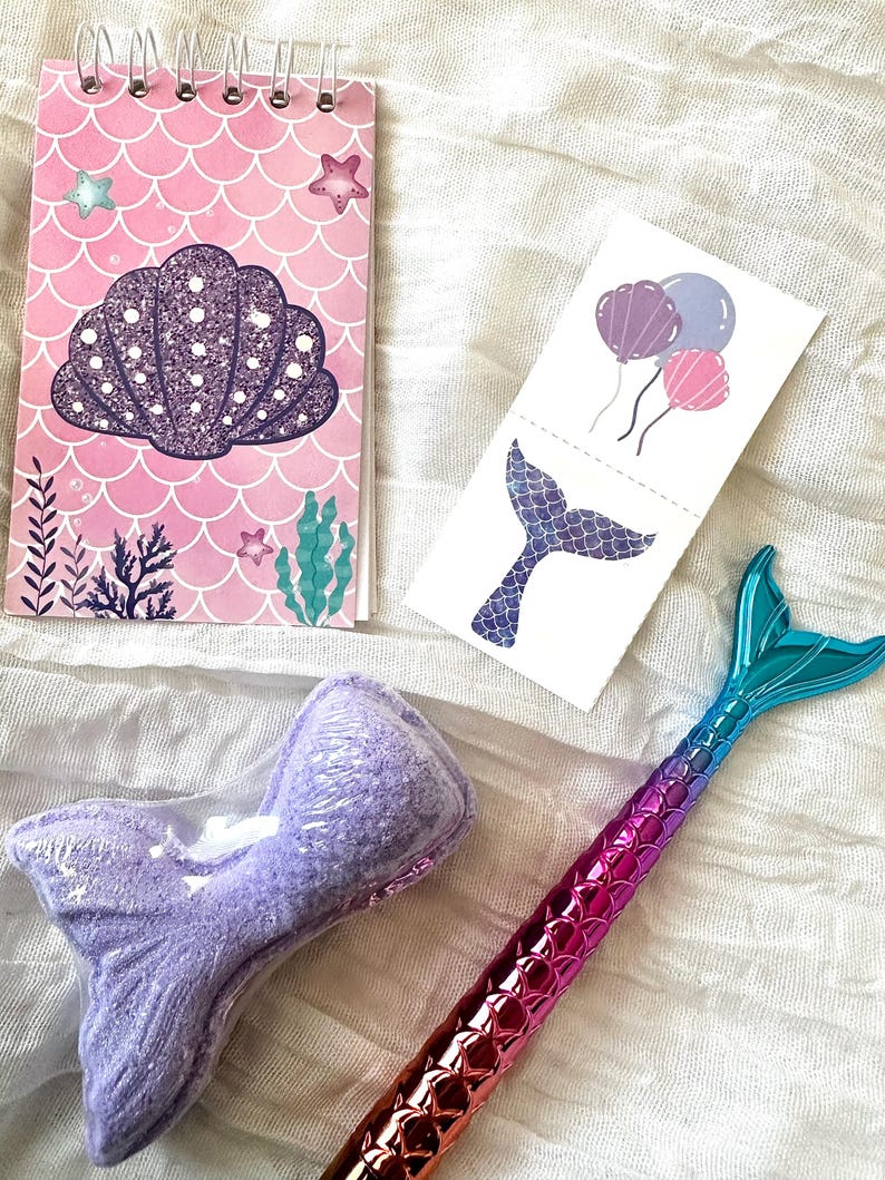 May include: A collection of mermaid-themed items, including a pink spiral notebook with a seashell design, temporary tattoos with balloons and a mermaid tail, a purple bath bomb, and a pen with a mermaid tail design.