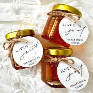 May include: Three small glass jars filled with honey, each with a gold lid and tied with twine. White tags with the text "LOVE IS sweet" and "MEGAN'S BRIDAL SHOWER" are attached to each jar. The jars are arranged on a white, textured surface.