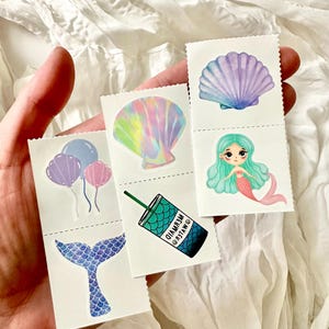 May include: A hand holds several small, rectangular stickers with mermaid-themed illustrations. The stickers feature a mermaid tail, balloons, a seashell, a mermaid, and a drink with the text "MYLER MERMAID". The stickers have a white background.
