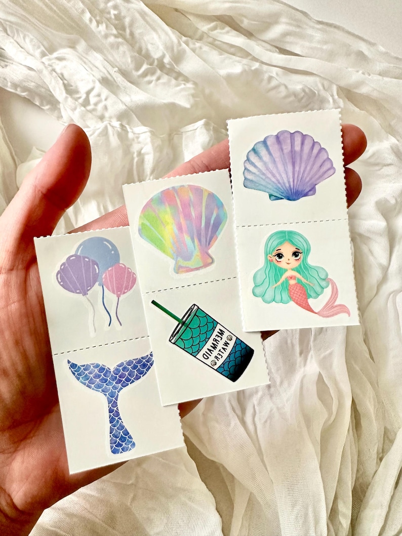 May include: A hand holds several small, rectangular stickers with mermaid-themed illustrations. The stickers feature a mermaid tail, balloons, a seashell, a mermaid, and a drink with the text "MYLER MERMAID". The stickers have a white background.