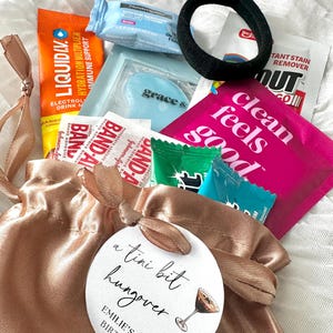 May include: A satin drawstring bag in a light brown color, tied with a matching ribbon, containing various small items. Includes packets of Liquid IV, Band-Aids, makeup remover, and a black hair tie. A round tag reads "a tini bit hungover".
