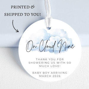 Pode incluir: Uma etiqueta redonda branca com um design de nuvem azul em aquarela e a frase "On Cloud Nine" em escrita cursiva. A etiqueta inclui o texto "THANK YOU FOR SHOWERING US WITH SO MUCH LOVE! BABY BOY ARRIVING MARCH 2026". Uma corda branca está anexada.