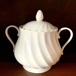 May include: A white porcelain sugar bowl featuring a swirling ribbed pattern on the body and lid. The container has two curved side handles and a small, pointed finial on top of the lid. The surface is smooth, glossy, and solid white.