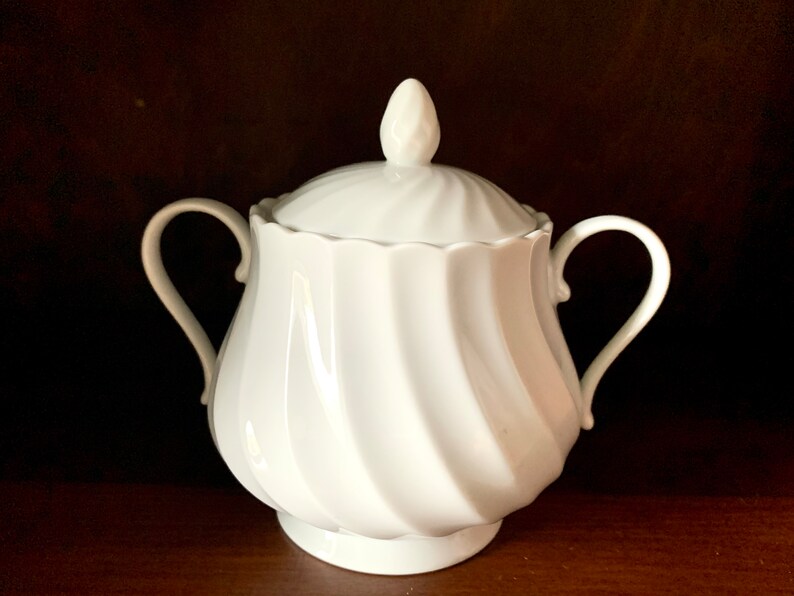 May include: A white porcelain sugar bowl featuring a swirling ribbed pattern on the body and lid. The container has two curved side handles and a small, pointed finial on top of the lid. The surface is smooth, glossy, and solid white.