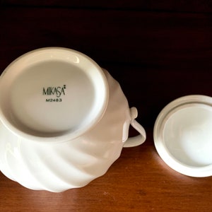 May include: A white porcelain sugar bowl with a swirling ribbed texture and two side handles. The base features the black text MIKASA and M2483. A matching circular lid with a smooth finish is placed beside the bowl on a dark wooden surface.