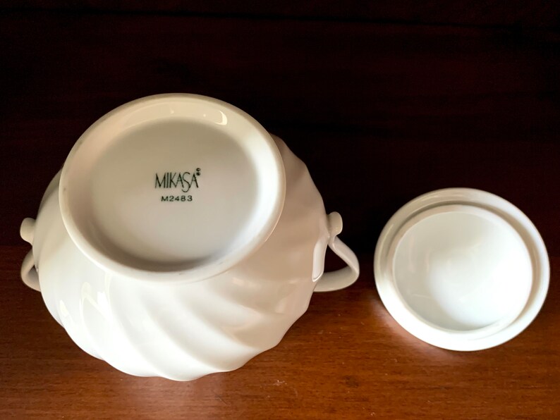 May include: A white porcelain sugar bowl with a swirling ribbed texture and two side handles. The base features the black text MIKASA and M2483. A matching circular lid with a smooth finish is placed beside the bowl on a dark wooden surface.