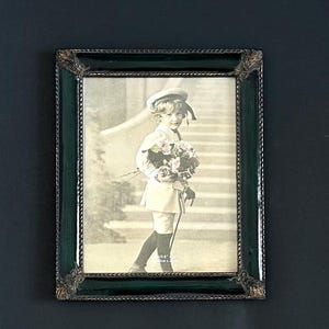 May include: A rectangular picture frame with a dark green, glossy border and ornate, metallic corner accents. The frame contains a sepia-toned photograph of a child in a cap and light-colored outfit holding a bouquet of flowers on stairs.