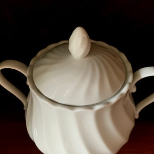 May include: A white porcelain sugar bowl featuring a swirling, fluted pattern on both the body and the lid. The lid is topped with a small, pointed finial, and the bowl includes two curved handles extending from opposite sides of the vessel.