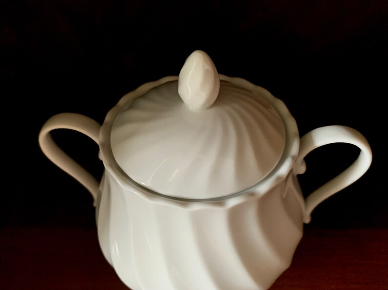 May include: A white porcelain sugar bowl featuring a swirling, fluted pattern on both the body and the lid. The lid is topped with a small, pointed finial, and the bowl includes two curved handles extending from opposite sides of the vessel.