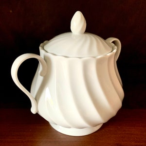 May include: A white porcelain sugar bowl featuring a swirling, ribbed pattern on the body and lid. The container has two curved side handles and a pointed finial on top of the lid. The surface is smooth, glossy, and uniform in color throughout.