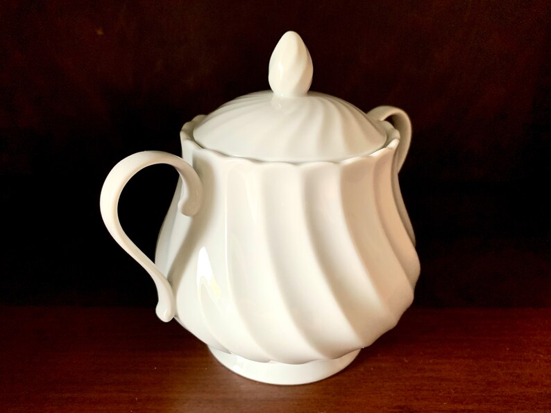 May include: A white porcelain sugar bowl featuring a swirling, ribbed pattern on the body and lid. The container has two curved side handles and a pointed finial on top of the lid. The surface is smooth, glossy, and uniform in color throughout.
