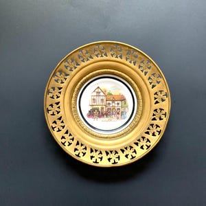 May include: A decorative plate featuring a central circular illustration of a timber-framed building with figures and a horse-drawn carriage, surrounded by a wide, ornate gold-toned metal border with a repeating cutout cross-like pattern.