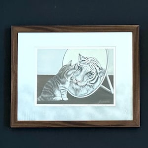 May include: A framed black and white art print featuring a small tabby kitten sitting in front of a round mirror. The reflection in the mirror shows the face of a large tiger. The artwork is enclosed in a simple brown wooden frame with a white mat.