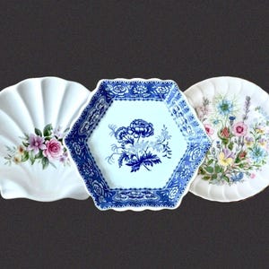 May include: Three ceramic plates: a white shell-shaped plate with a pink rose floral design, a hexagonal plate with a blue and white floral pattern, and a round plate with a colorful botanical illustration of various wildflowers and greenery.