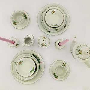 May include: A white porcelain tea set with a green holly design. The set includes a teapot, creamer, sugar bowl, two cups and saucers, and two plates. There are also two pink candles and candle holders.