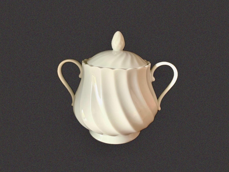 May include: A white porcelain sugar bowl featuring a swirling, ribbed pattern across its rounded body. The vessel includes two curved side handles, a matching lid with a pointed finial, and a scalloped rim edge, all finished in a glossy glaze.