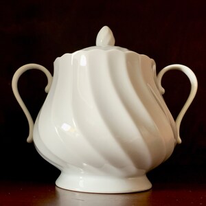 May include: A white porcelain sugar bowl featuring a distinct spiral swirl pattern across its rounded body. The vessel includes two curved side handles, a matching lid with a pointed finial, and a scalloped rim edge, all finished in a glossy glaze.
