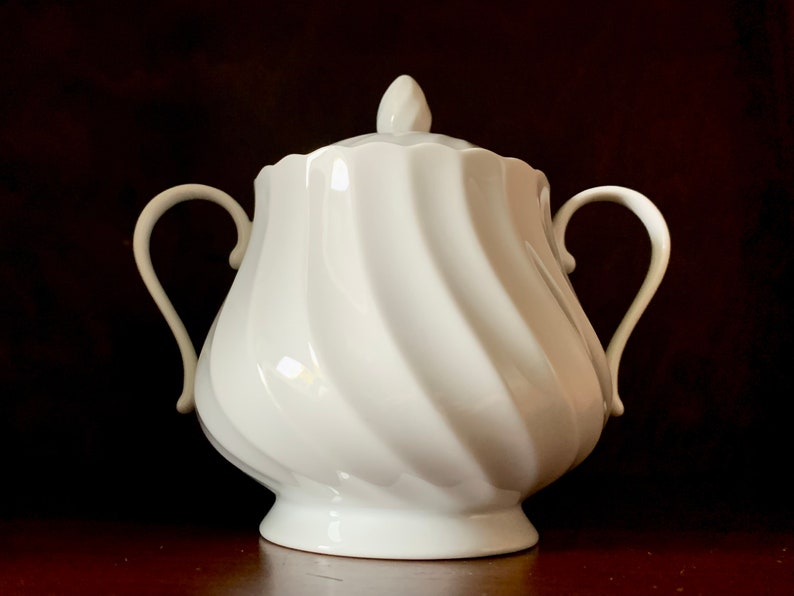 May include: A white porcelain sugar bowl featuring a distinct spiral swirl pattern across its rounded body. The vessel includes two curved side handles, a matching lid with a pointed finial, and a scalloped rim edge, all finished in a glossy glaze.