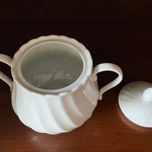 May include: A white porcelain sugar bowl featuring a scalloped rim and a swirling, ribbed texture on the body. The bowl includes two curved side handles and a matching circular lid with a small knob handle, both displaying a consistent glossy finish.