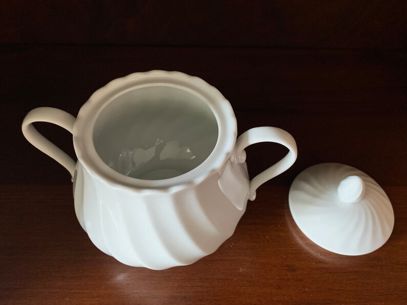 May include: A white porcelain sugar bowl featuring a scalloped rim and a swirling, ribbed texture on the body. The bowl includes two curved side handles and a matching circular lid with a small knob handle, both displaying a consistent glossy finish.